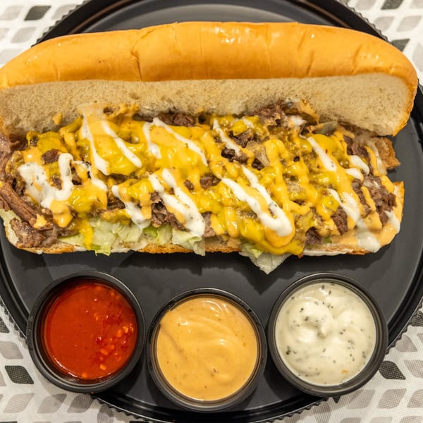 Cheesesteak hoagie with chopped beef, melted cheese, lettuce, and drizzled sauces, served with three dipping sauces on a black plate.
