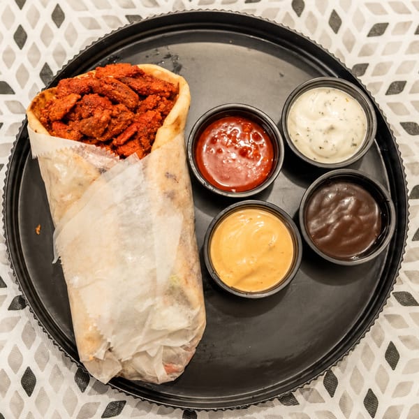 A wrap filled with crispy red-seasoned chicken sits on a black platter beside four dipping sauces.