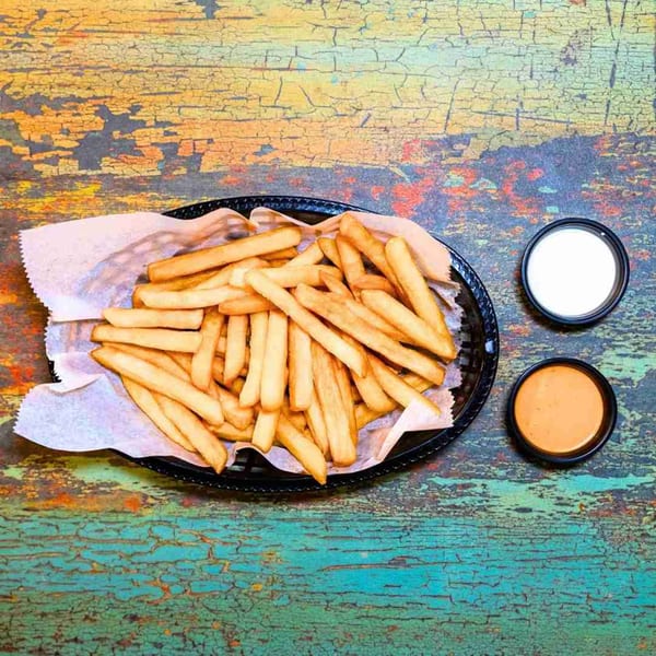 A basket of golden french fries with two dipping sauces, one white and one orange, on a distressed wooden table.