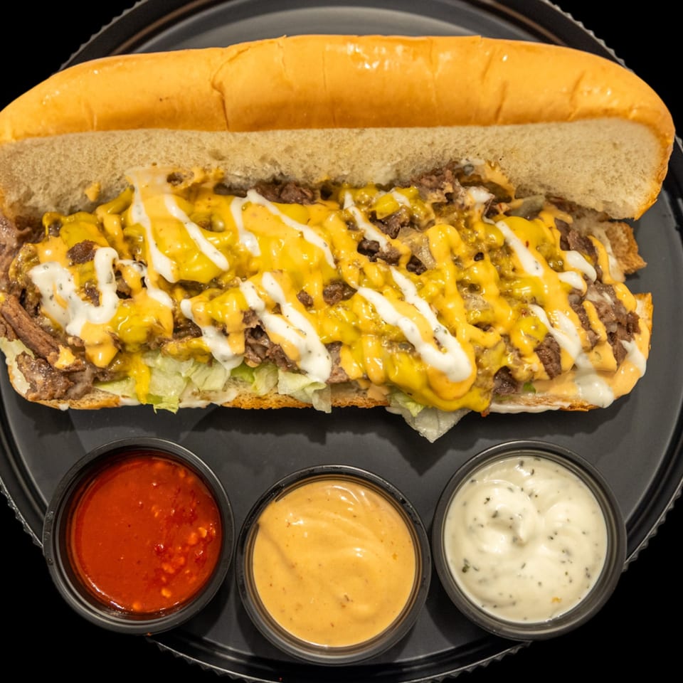 Loaded cheesesteak sandwich with chopped beef, melted cheese, lettuce, and three dipping sauces on a black plate.