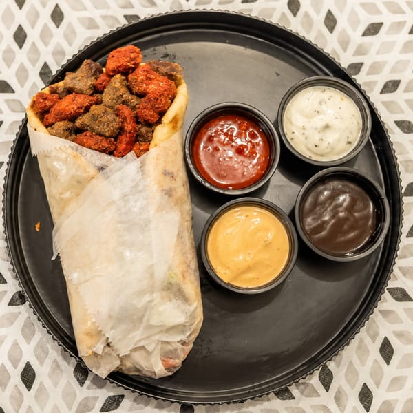 Wrapped flatbread filled with grilled meat pieces beside four dipping sauces on a black serving tray.