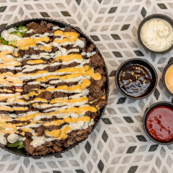Platter of grilled meat topped with yellow and white sauces, accompanied by assorted dipping bowls on a patterned tablecloth.