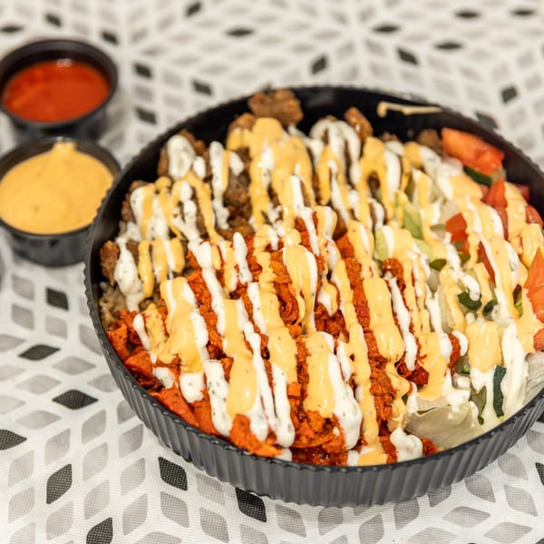 Loaded platter of seasoned fries topped with shredded meat, vegetables, and drizzled white and orange sauces, with dipping sauces alongside.