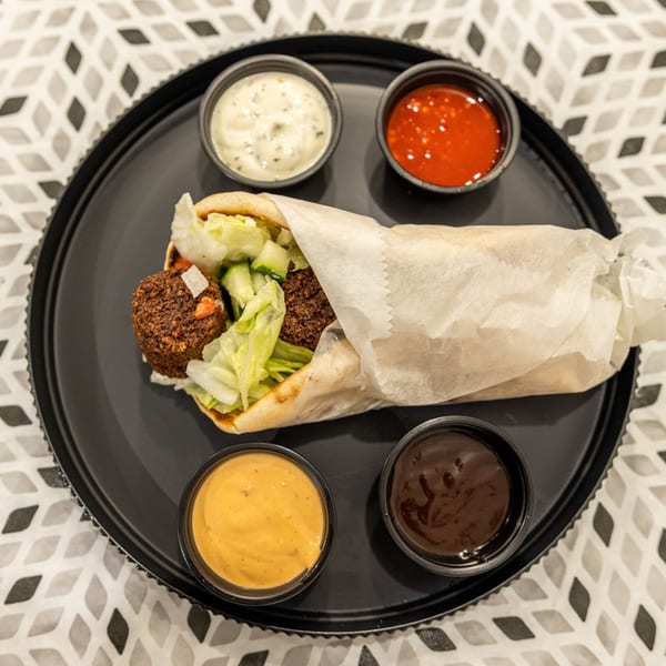 Falafel pita stuffed with lettuce and vegetables, served on a black tray with four dipping sauces.