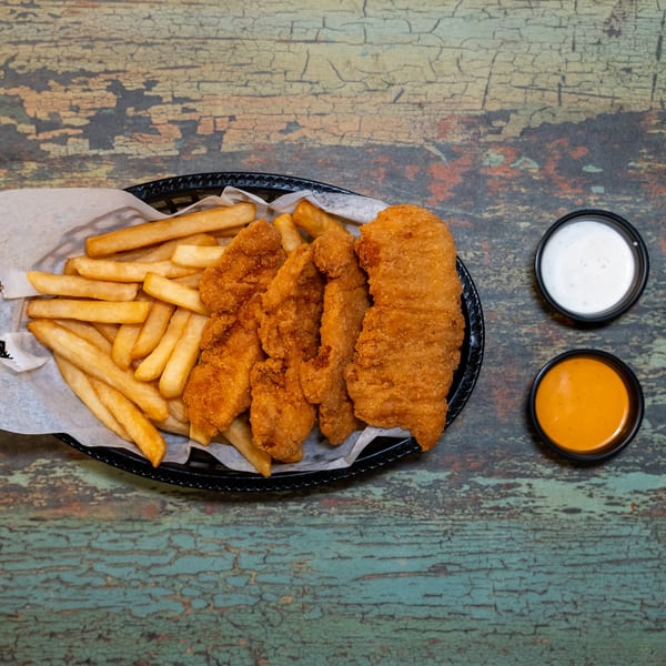 A basket of golden-brown chicken tenders and french fries with two dipping sauces on a rustic table.