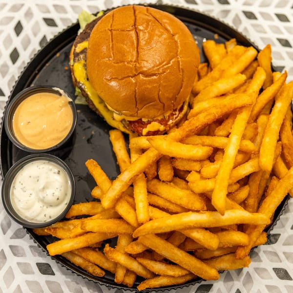 Cheeseburger with a glossy bun, golden French fries, and two dipping sauces on a black plate.