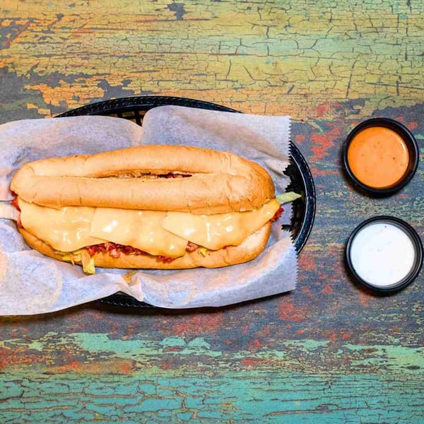 A DC BITES halal cheesesteak sandwich in a basket with two dipping sauces on a distressed wooden table.