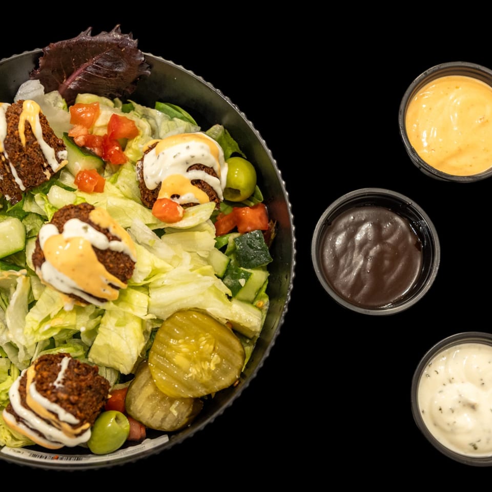 Salad bowl with falafel, lettuce, tomatoes, cucumbers, pickles, olives, and drizzled sauces, alongside three dipping sauces.